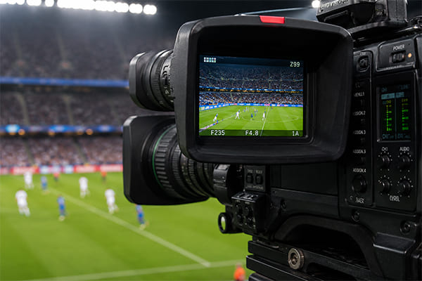 A close-up of a broadcast camera filming a soccer match under bright, uniform lights